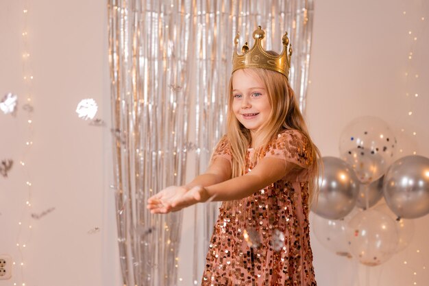 una niña feliz con un vestido elegante y una corona sopla confetti de sus palmas y pide un deseo.