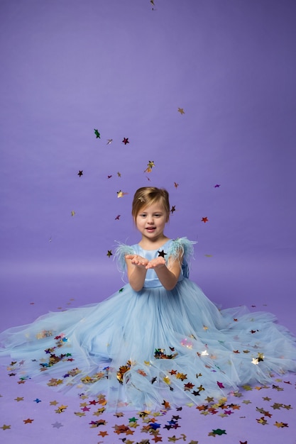 Una niña feliz con un vestido de princesa se sienta y atrapa confeti con las manos en púrpura