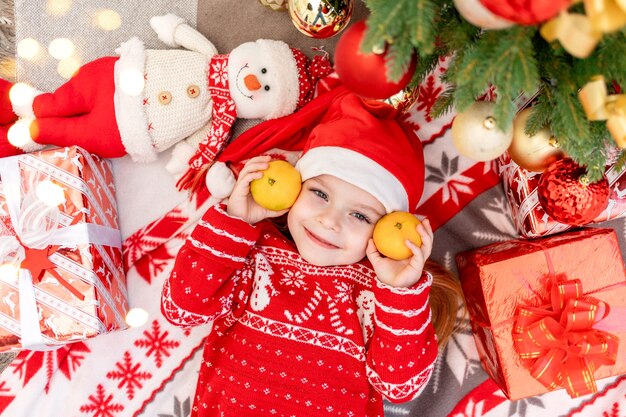 Una niña feliz yace bajo el árbol de Navidad en casa con mandarinas y espera el año nuevo o la Navidad con un sombrero rojo de Santa Claus y sonríe y se regocija