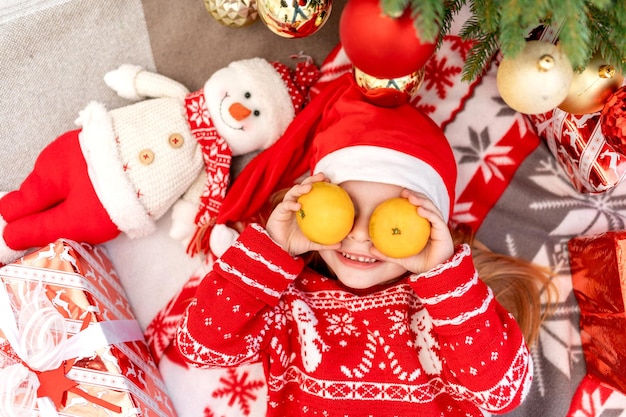 Una niña feliz yace bajo el árbol de Navidad en casa con mandarinas y espera el año nuevo o la Navidad con un sombrero rojo de Santa Claus y sonríe y se regocija
