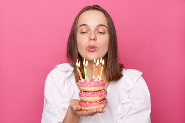 Niña festiva de cabello castaño sosteniendo deliciosos donuts apetitosos y soplando velas pidiendo deseos viste blusa blanca celebrando cumpleaños aislada sobre fondo rosa