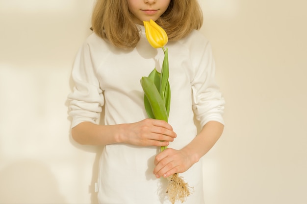 Niña con flor de tulipán amarillo, soleado fondo de pared brillante, primer plano