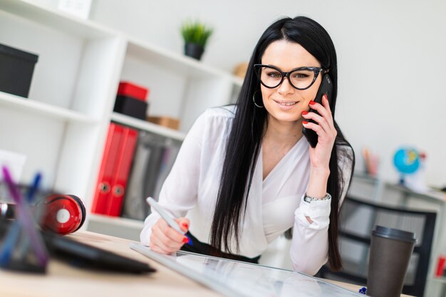 Una niña con gafas se para cerca de una mesa, habla por teléfono y dibuja un marcador en una pizarra magnética.