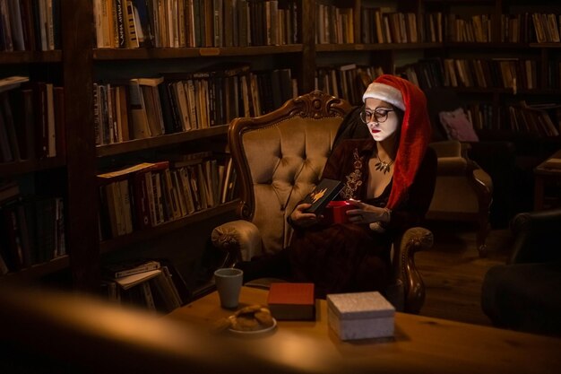 Niña con gafas en gorro de Papá Noel abre un regalo de Navidad sentado en un sillón en una biblioteca