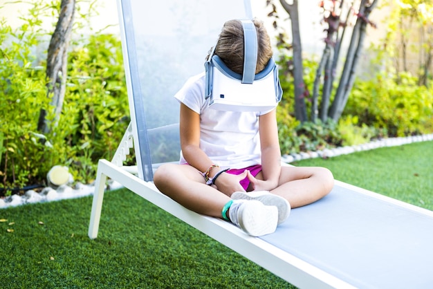 Niña con gafas de realidad virtual mirando hacia abajo sentada en una tumbona en el jardín de su casa Metaverse VR juego digital y concepto de simulación