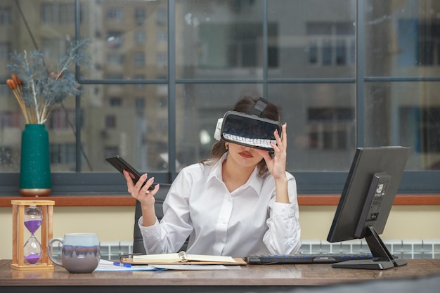 Niña con gafas de realidad virtual y sosteniendo su teléfono mientras está sentada en el escritorio