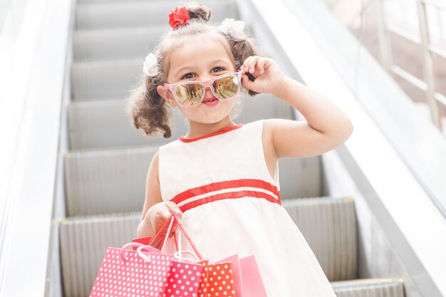 Niña con gafas de sol en la escalera mecánica del centro comercial con compras