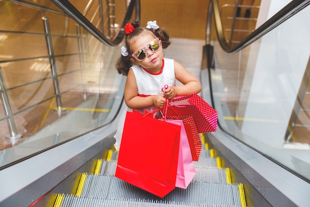 Niña con gafas de sol en la escalera mecánica del centro comercial con compras