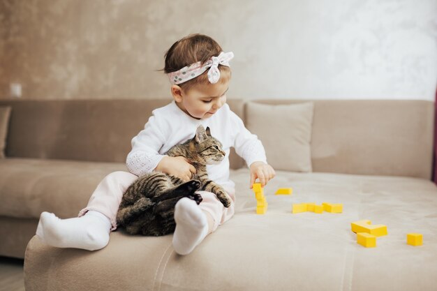 niña con gatito atigrado sentado en el sofá y juega con cubos amarillos