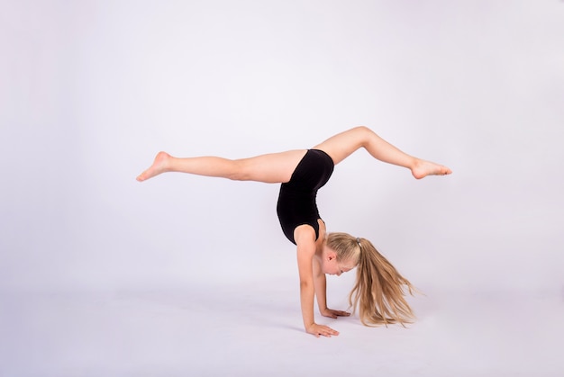 Una niña gimnasta en traje de baño negro realiza el pino sobre una pared blanca aislada