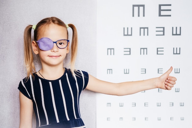 Niña con glasse y parche en el ojo u oclusor con el pulgar hacia arriba y tabla optométrica borrosa en el fondo, tratamiento de ambliopía (ojo vago)