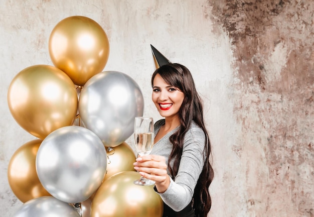 Una niña con globos se ríe contra el fondo de una pared decorativa Una hermosa mujer feliz en el cumpleaños de una fiesta de Año Nuevo divirtiéndose sosteniendo una copa de champán