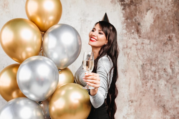 Una niña con globos se ríe contra el fondo de una pared decorativa Una hermosa mujer feliz en el cumpleaños de una fiesta de Año Nuevo divirtiéndose sosteniendo una copa de champán