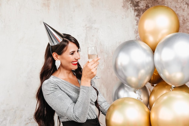 Una niña con globos se ríe contra el fondo de una pared decorativa Una hermosa mujer feliz en el cumpleaños de una fiesta de Año Nuevo divirtiéndose sosteniendo una copa de champán