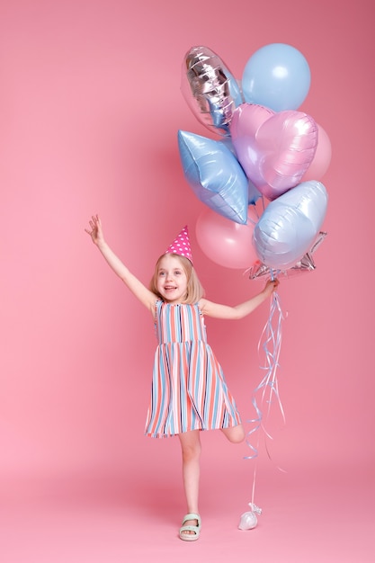 Niña con una gorra en la cabeza con globos sobre una superficie rosa