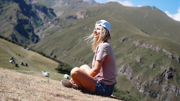 La niña con gorra y pantalones cortos en las montañas se sienta en la hierba