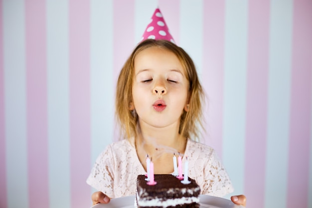 Niña con gorra rosa soplando velas en un pastel de chocolate de cumpleaños en su fiesta de cumpleaños en casa. Retrato cumpleaños niño.