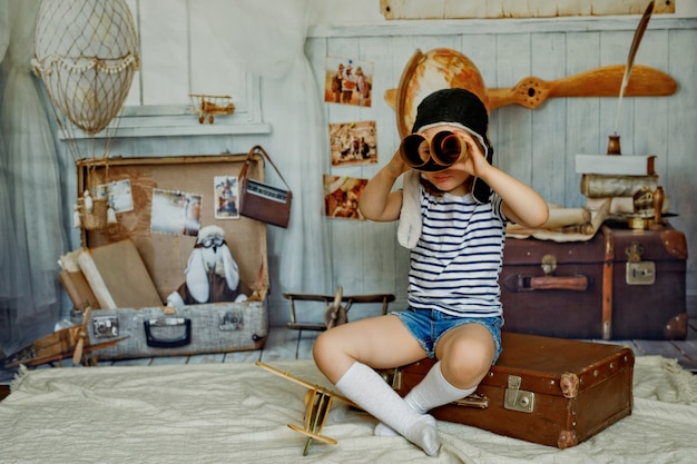 Una niña con una gorra se sienta en una maleta retro y sostiene un mapa en su mano como un telescopio