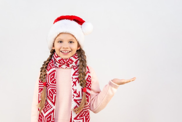 Una niña con un gorro y bufanda navideños señala con la palma hacia un lado y sonríe.