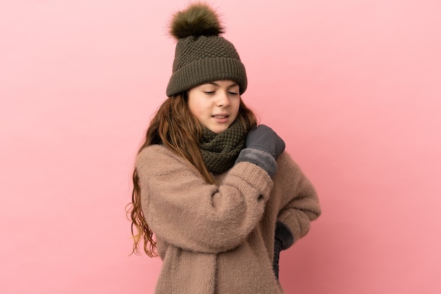 Niña con gorro de invierno aislado sobre fondo rosa que sufre de dolor en el hombro por haber hecho un esfuerzo