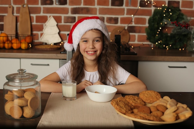 Una niña con un gorro de Papá Noel en la cocina junto a un tarro de galletas y un vaso de leche