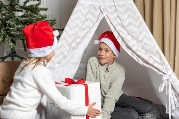 Una niña con un gorro de Papá Noel le da un regalo a un niño por Navidad y Año Nuevo Hermano y hermana siguen intercambiando regalos