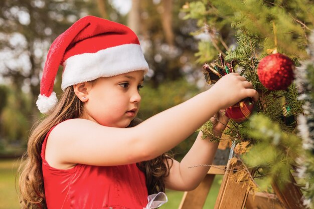 Niña con gorro de Papá Noel decorando el árbol de Navidad.