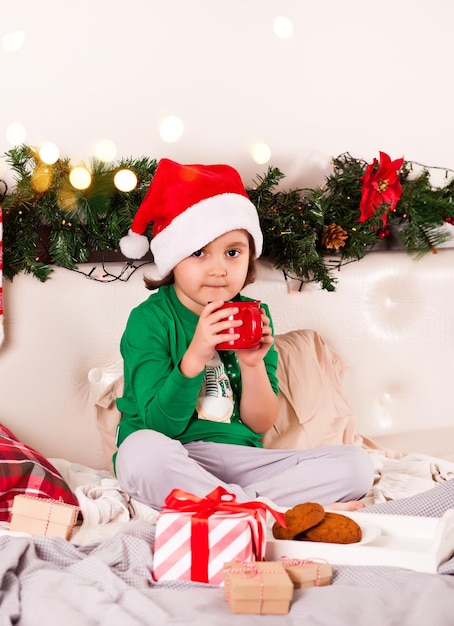 Niña con gorro de Papá Noel divertido y pijama de Navidad sentado en una cama comiendo galletas y bebiendo cacao