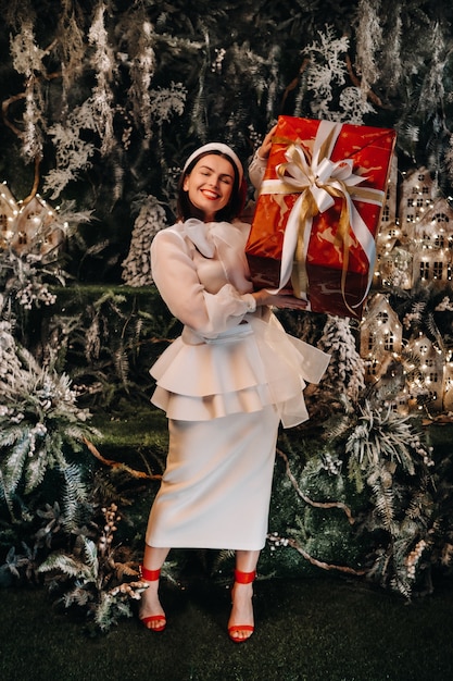 Una niña con un gorro de Papá Noel con un gran regalo de Navidad en sus manos en una isla de cuento de hadas. Mujer sonriente en ropa blanca en el fondo de árboles de Navidad y casas pequeñas.