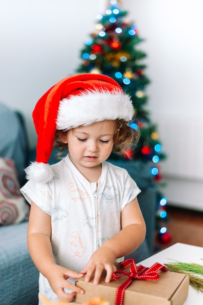 Niña con gorro de Papá Noel y pijama abriendo regalos junto al árbol de Navidad con luces de hadas