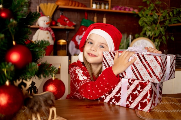 Una niña con un gorro de Papá Noel con regalos en una cocina oscura junto a un árbol de Navidad con bolas rojas se regocija y sonríe, el concepto de año nuevo y Navidad