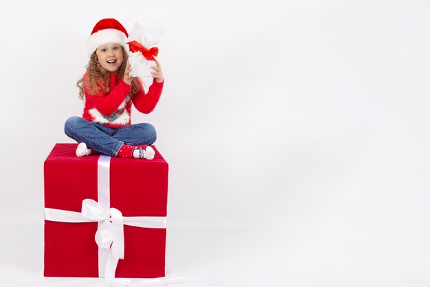 Niña con un gorro de Papá Noel se sienta en un cubo rojo con un regalo en sus manos infancia feliz