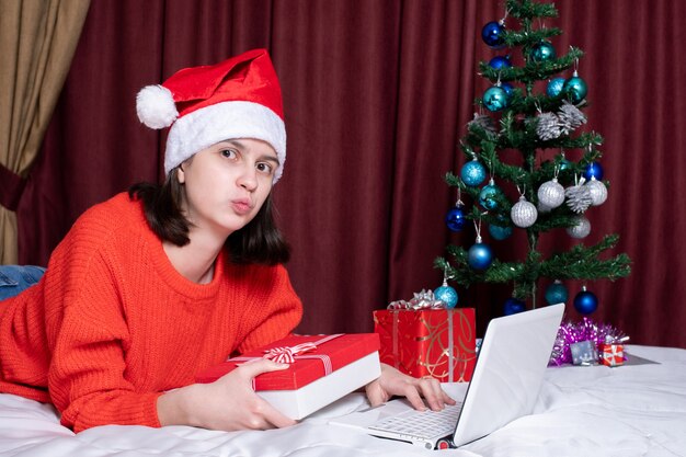 Una niña con un gorro de Papá Noel y un suéter de punto rojo pasa un beso para regalos o compras. Concepto de felicitaciones navideñas durante el autoaislamiento. Regalos y compras navideñas