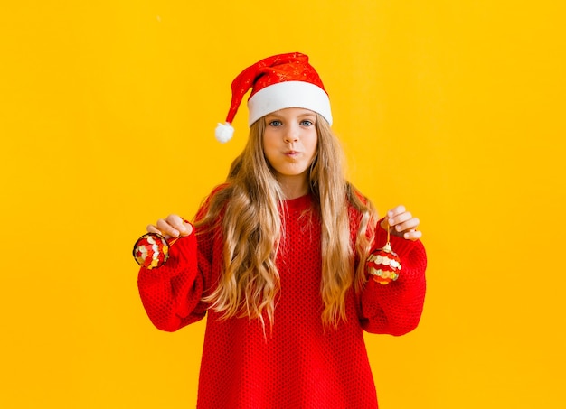 Niña con un gorro de Papá Noel y un suéter rojo salta y muestra cinco sobre un fondo amarillo.