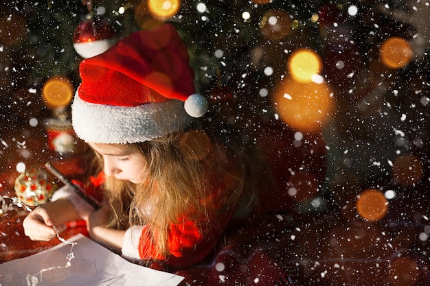 Niña con un gorro de Papá Noel y un vestido rojo debajo del árbol de Navidad está soñando, esperando las vacaciones, acostada sobre una manta a cuadros. Una carta en papel, regalos. Año nuevo, Navidad. luces de desenfoque