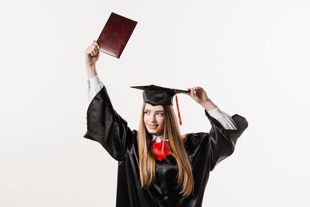 La niña graduada se gradúa de la universidad y celebra los logros académicos Una estudiante feliz con un vestido de graduación negro y una gorra levanta el diploma de maestría por encima de la cabeza sobre fondo blanco