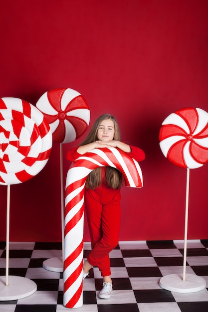 Niña con un gran bastón de caramelo de Navidad en una pared roja aislada. Enormes dulces navideños. dulces