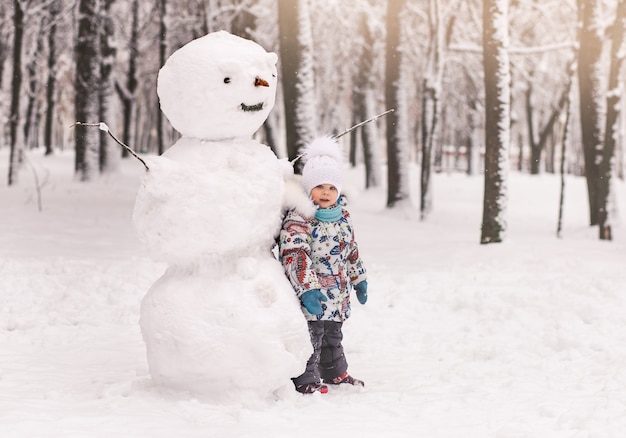 Niña y un gran muñeco de nieve de invierno en el parque