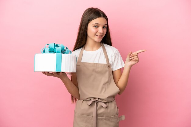 Niña con un gran pastel sobre fondo rosa aislado que señala con el dedo hacia el lado