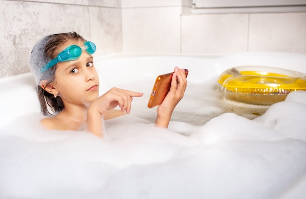 Niña hace un selfie con un teléfono inteligente mientras se baña en un baño de burbujas en casa