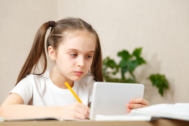 Niña haciendo los deberes en casa en la mesa. El niño es educado en casa. Una niña de cabello claro realiza una tarea en línea usando una computadora portátil y una tableta.