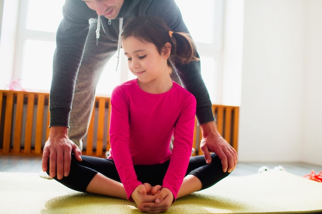 Foto niña está haciendo ejercicios de estiramiento entrenamiento en casa. lindo niño y papá están entrenando en una estera de interior. la pequeña modelo de cabello oscuro en ropa deportiva tiene ejercicios cerca de la ventana de su habitación