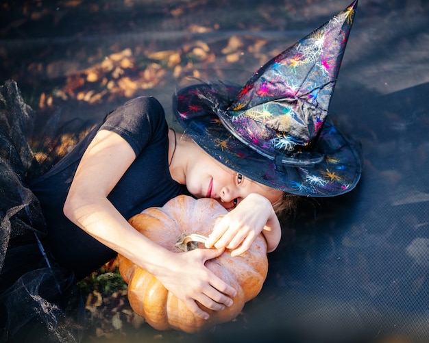 Niña de Halloween con sombrero de bruja sosteniendo una calabaza y acostado en el trampolín con hojas amarillas en el fondo