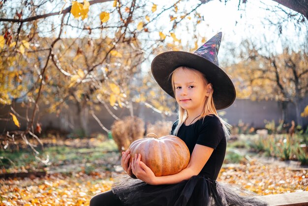 Niña de Halloween con sombrero de bruja sosteniendo una calabaza con hojas amarillas en el fondo