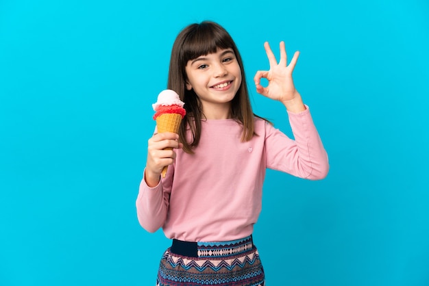 Niña con un helado de cucurucho aislado en la pared azul que muestra el signo de ok con los dedos