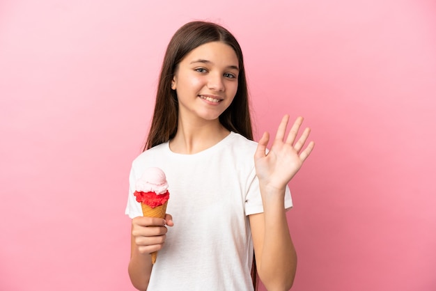 Niña con un helado de cucurucho sobre fondo rosa aislado saludando con la mano con expresión feliz