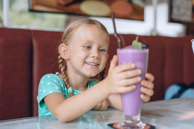 Una niña hermosa con coletas rubias está mirando un gran vaso de batido y disfrutando de un postre dulce en un acogedor café local. Retrato.