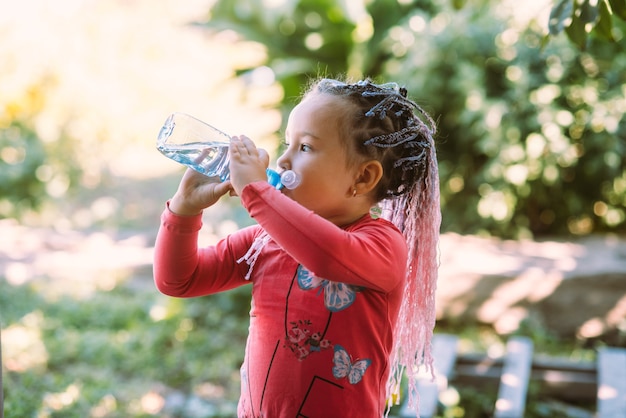 Una niña hermosa con coletas trenzadas bebe agua al aire libre en el jardín