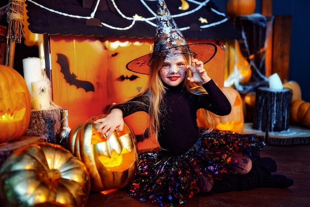 Una niña hermosa en un disfraz de bruja celebra en casa en un interior con calabazas y casa mágica de cartón en el fondo