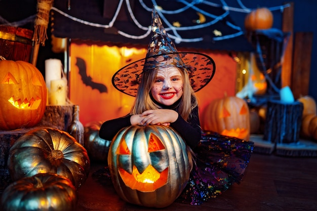 Una niña hermosa en un disfraz de bruja celebra en casa en un interior con calabazas y casa mágica de cartón en el fondo